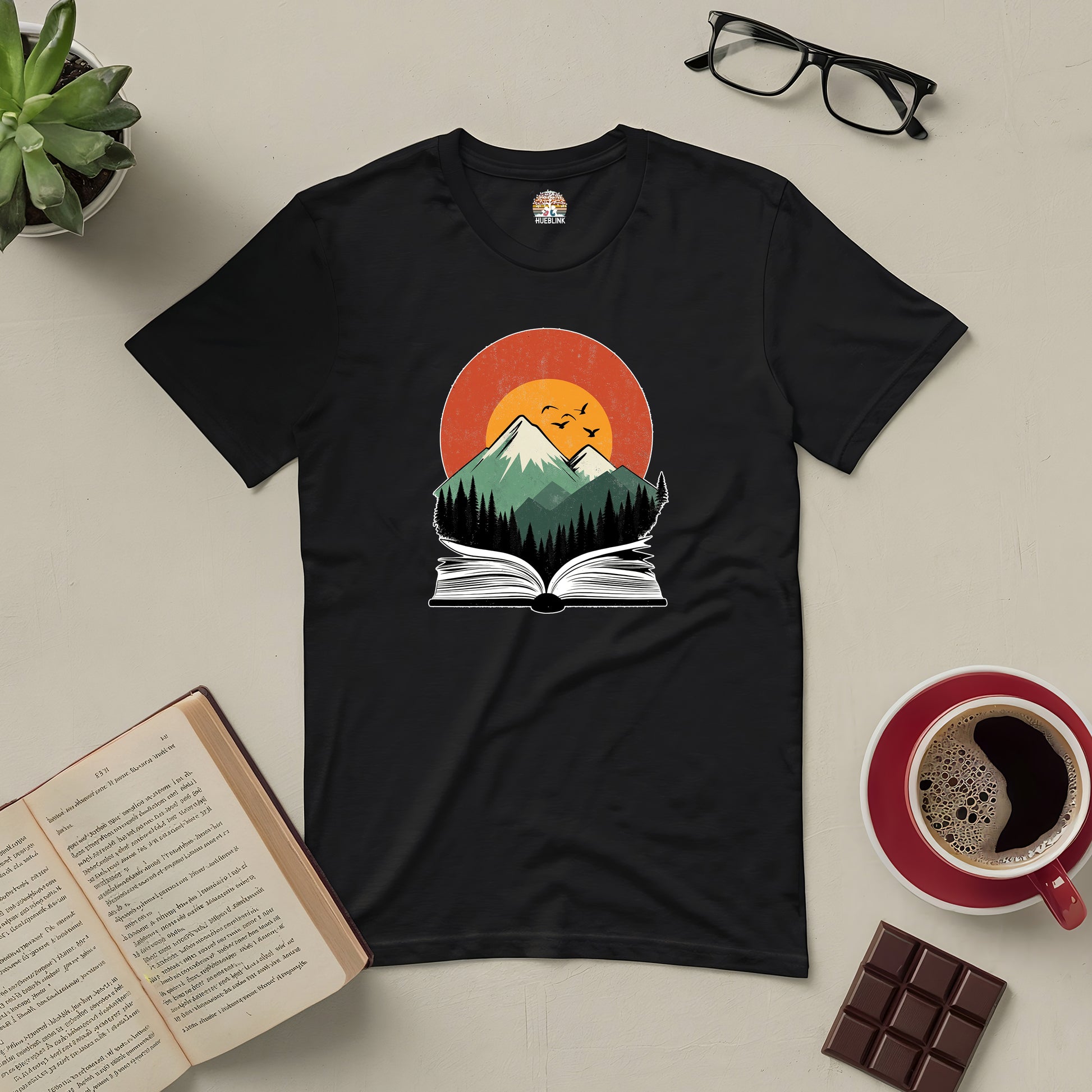 Black t-shirt with mountain and book design, surrounded by coffee, glasses, plant, and chocolate on a beige surface.