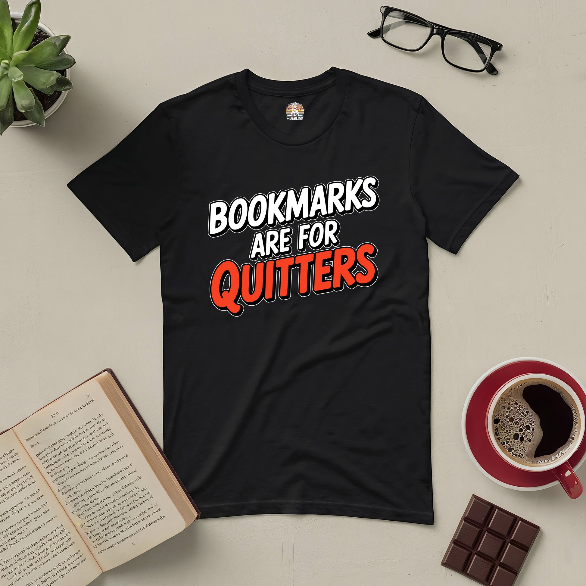 Black "Bookmarks Are for Quitters" T-shirt for avid readers, styled with glasses, book, coffee, plant, and chocolate on a light table.