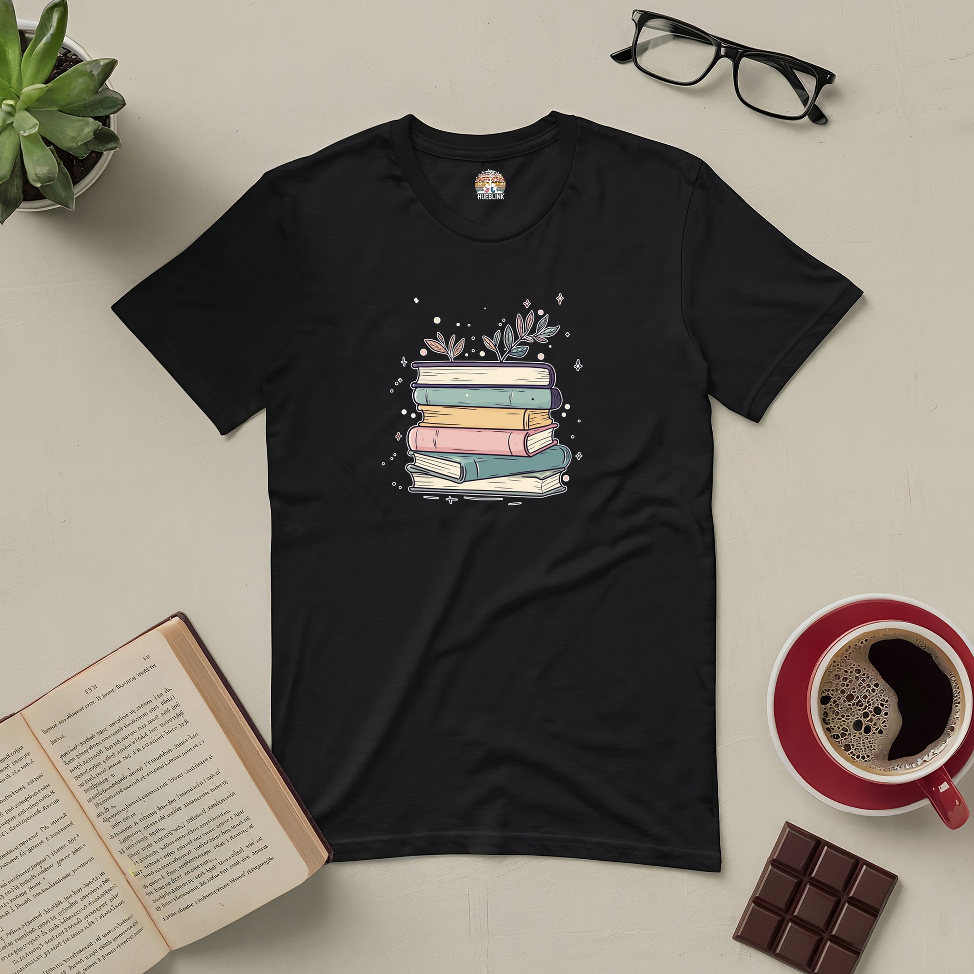 Black tee featuring pastel stack of books with floral accents, surrounded by coffee, glasses, chocolate, and an open book.