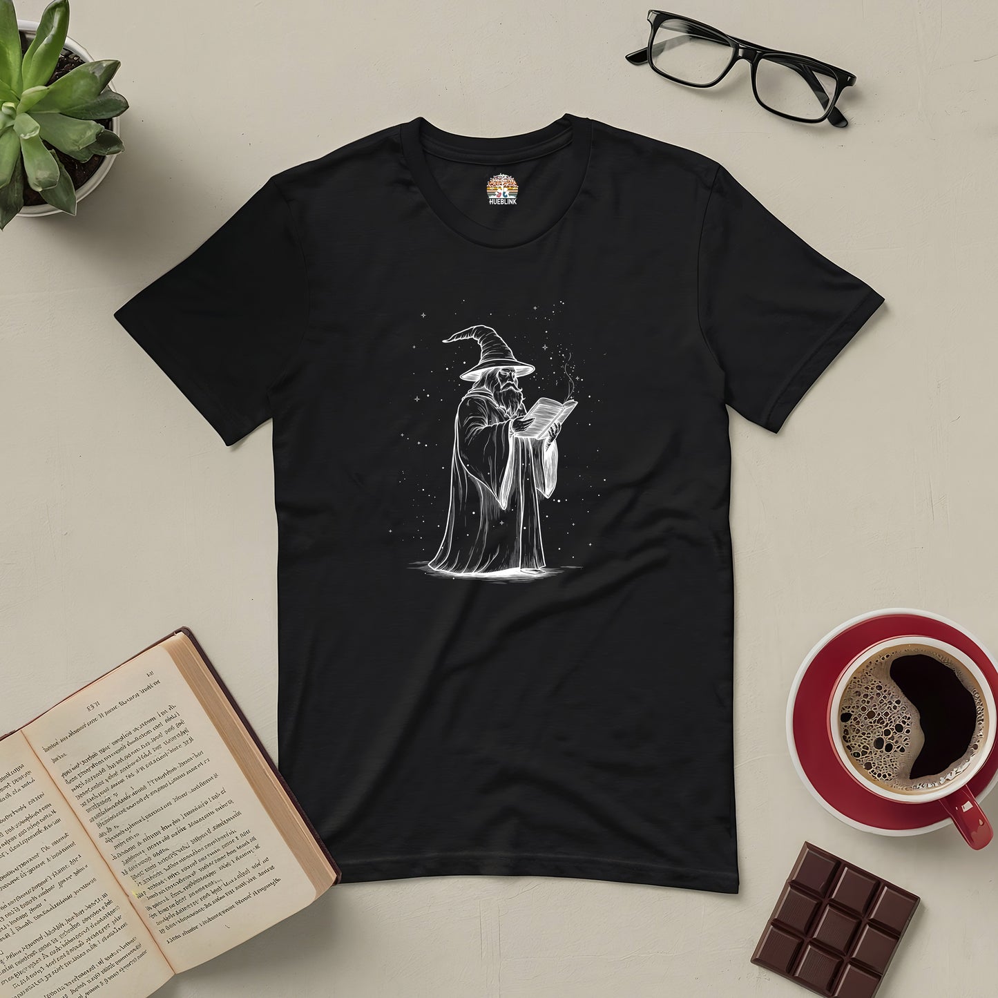 Wizard's Grimoire Tee with mystical wizard design, ideal for book lovers and fantasy fans, laid with book, coffee, and chocolate bar.