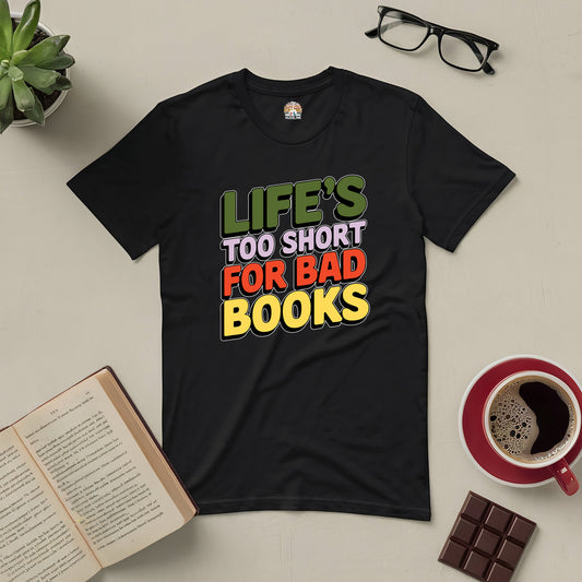 Black tee with "Life's Too Short for Bad Books" text, surrounded by glasses, coffee, chocolate, plant, and open book. Perfect for book lovers.