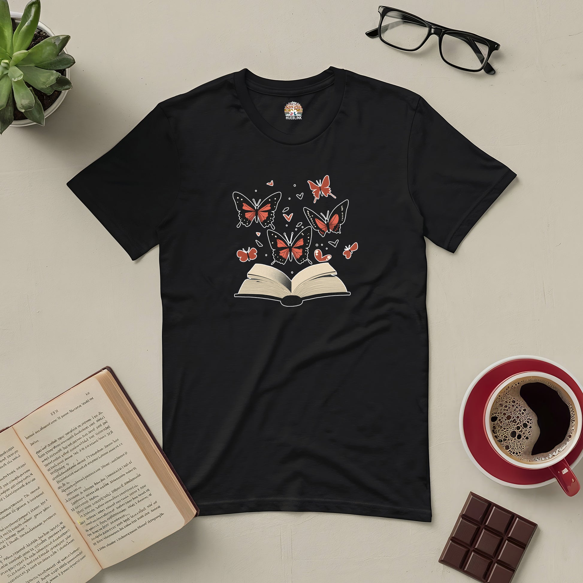 Black "Books: Where Stories Take Flight" tee with open book and butterfly design, beside coffee, chocolate, glasses, and an open book.