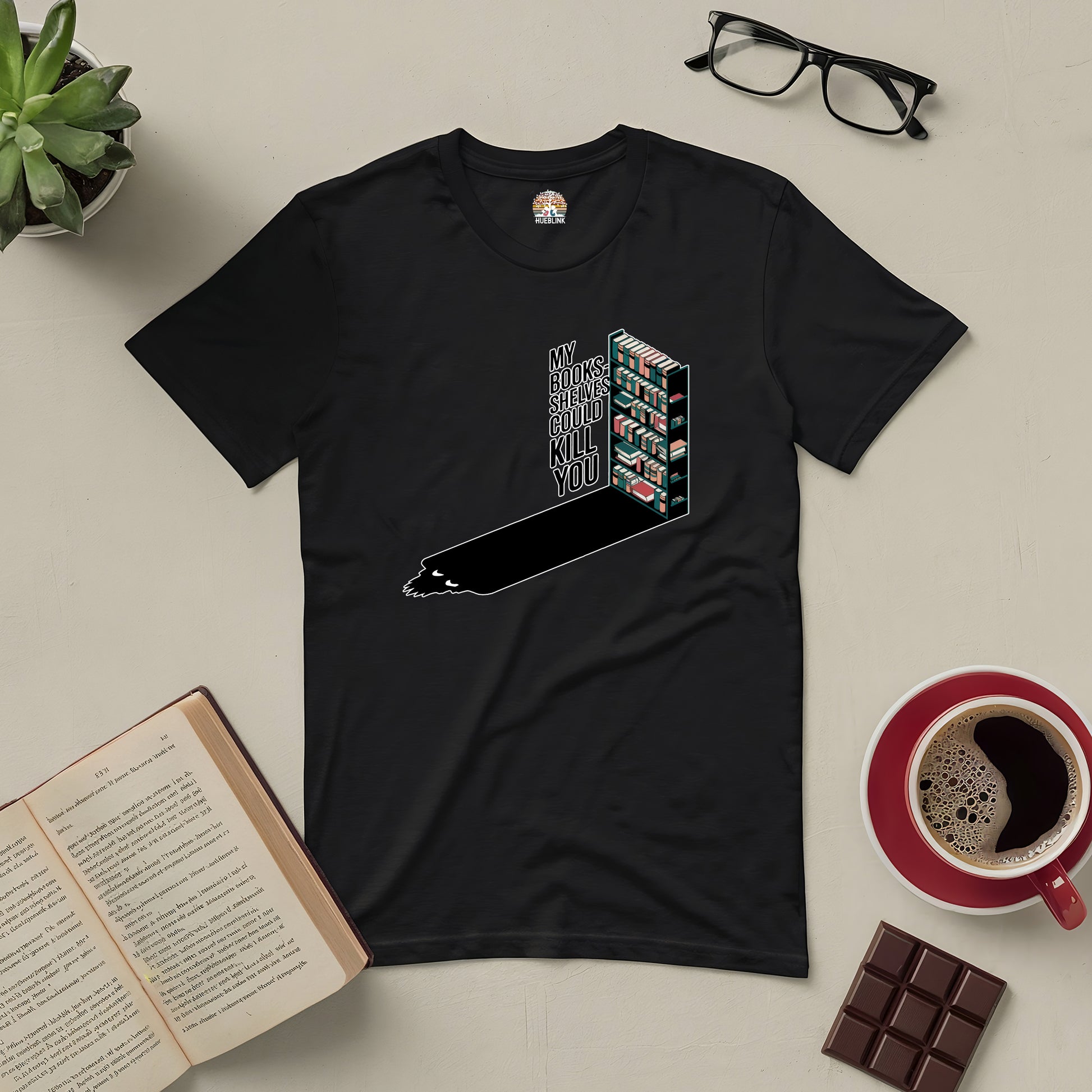 Black "My Bookshelves Could Kill You" tee for book lovers, surrounded by open book, glasses, coffee, chocolate, and plant.