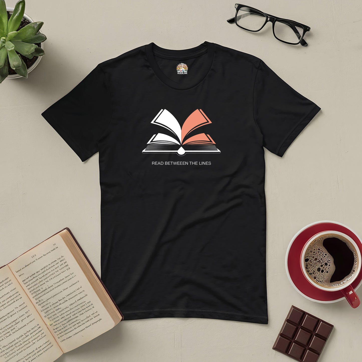 Black "Read Between The Lines" tee with open book design, surrounded by glasses, open book, coffee cup, chocolate, and a plant.