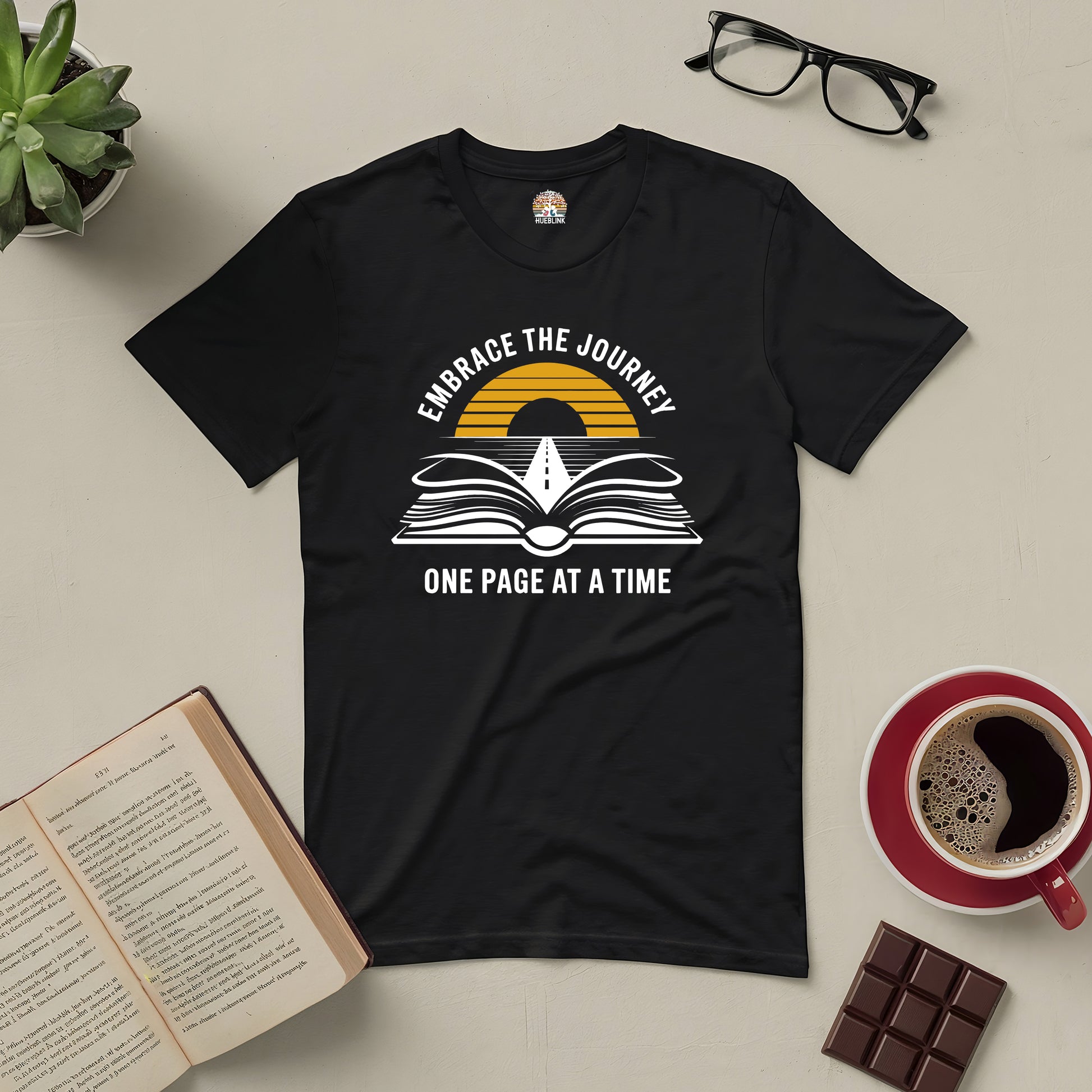 Black tee with "Embrace the Journey" design, featuring an open book and road leading to horizon. Inspirational apparel laid on a table.