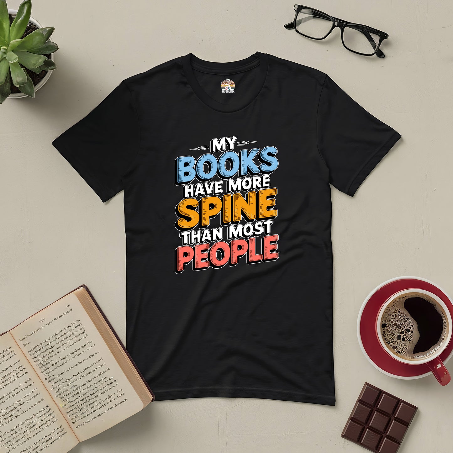 Black tee with "My Books Have More Spine Than Most People" text, surrounded by a book, coffee, chocolate, glasses, and a plant.