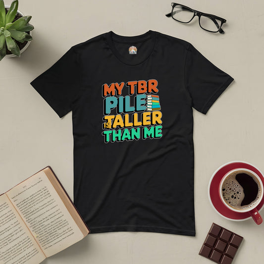 Black "My TBR Pile is Taller Than Me" tee with colorful text, surrounded by books, glasses, coffee, chocolate, and a plant.