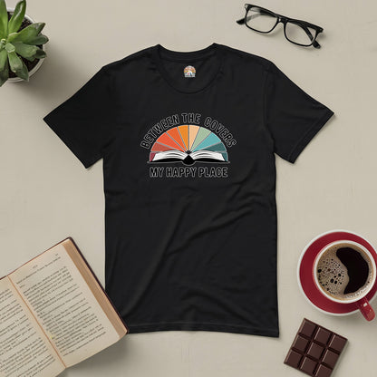 Black "Between The Covers: My Happy Place" tee with open book design, surrounded by glasses, coffee cup, book, chocolate, and plant.