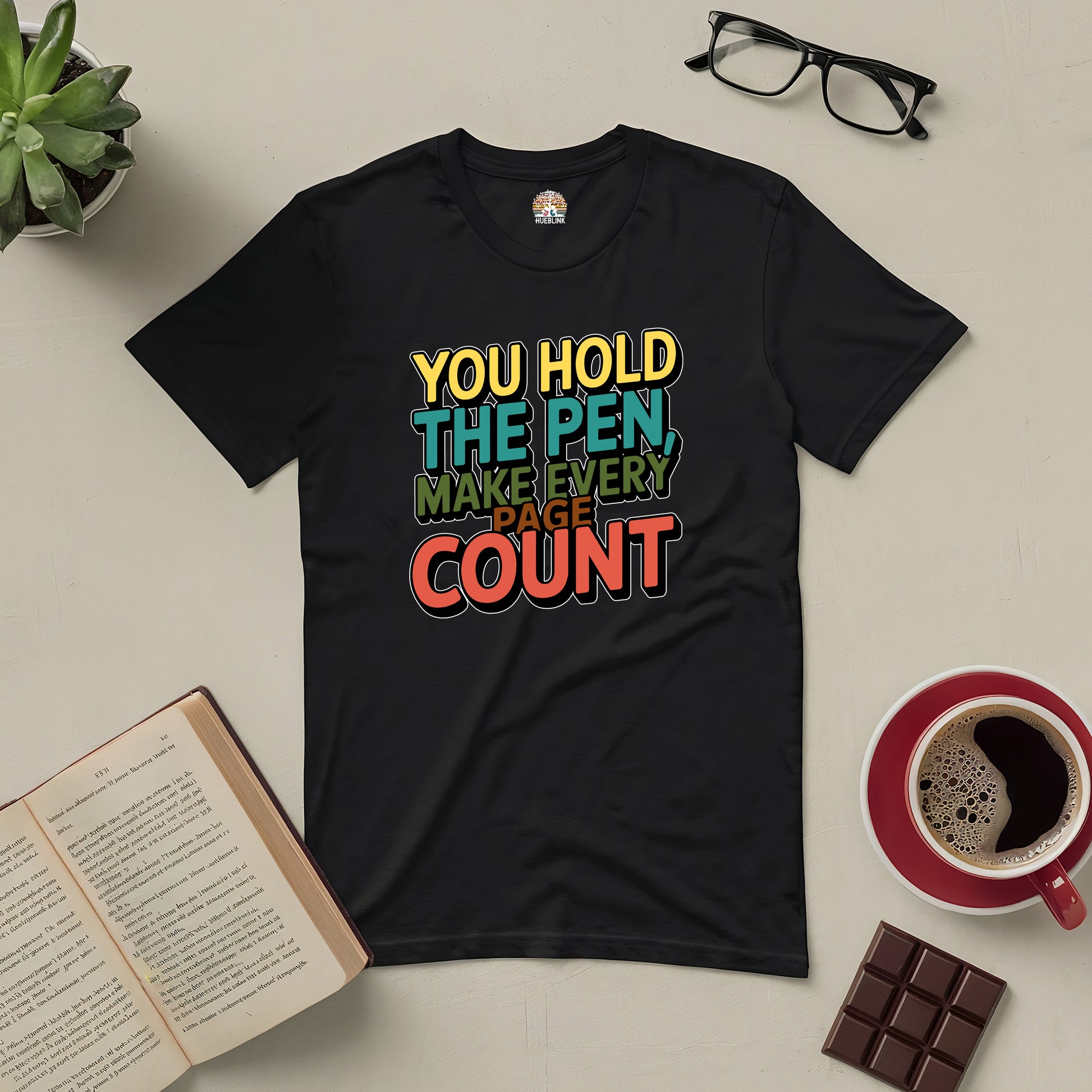 Black tee with "You Hold the Pen, Make Every Page Count" text, placed on a table with books, glasses, coffee, and chocolate.