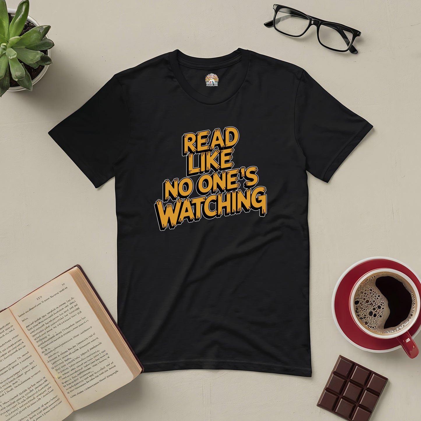 Black tee with "Read Like No One's Watching" text, surrounded by glasses, a book, coffee, chocolate, and a plant.