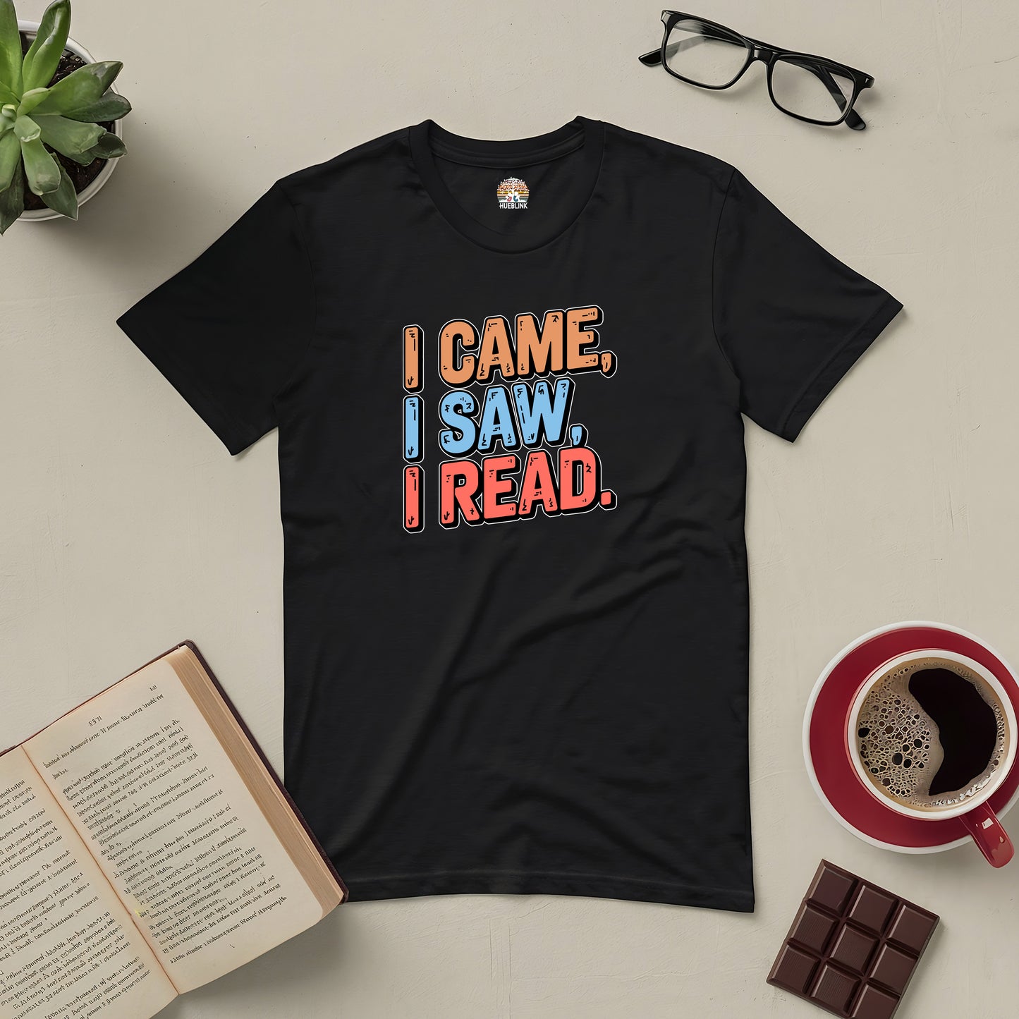 Black tee with "I Came, I Saw, I Read" design, surrounded by an open book, glasses, coffee, chocolate, and a plant. Perfect for book lovers.