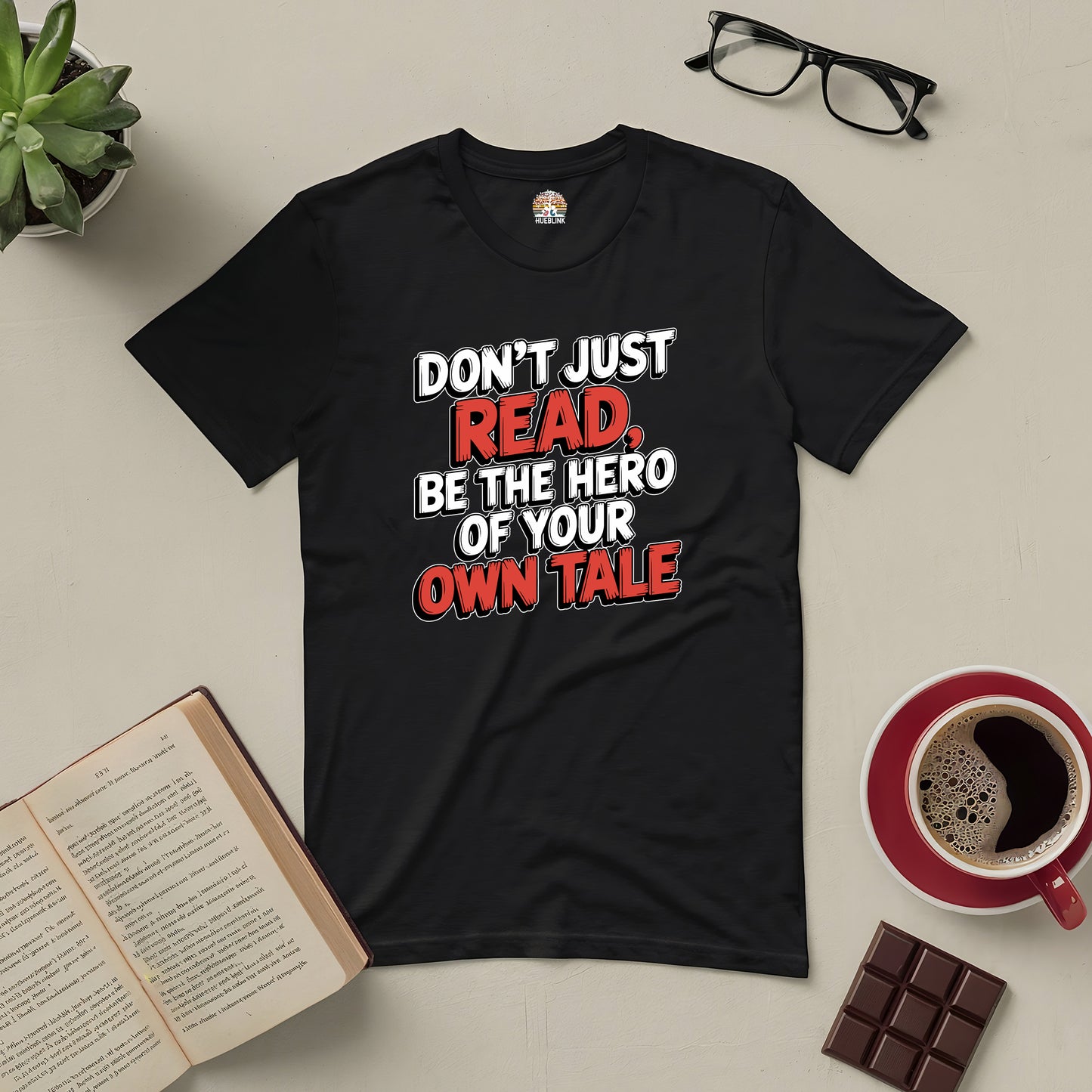 Black tee with "Don't Just Read, Be the Hero of Your Own Tale" text, surrounded by books, coffee, chocolate, and glasses.