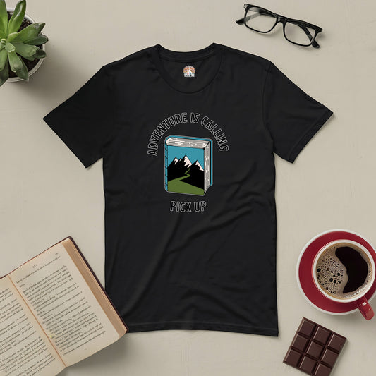 Black Adventure Is Calling Pick Up Tee with mountain graphic, surrounded by a book, glasses, chocolate, and coffee on a table.