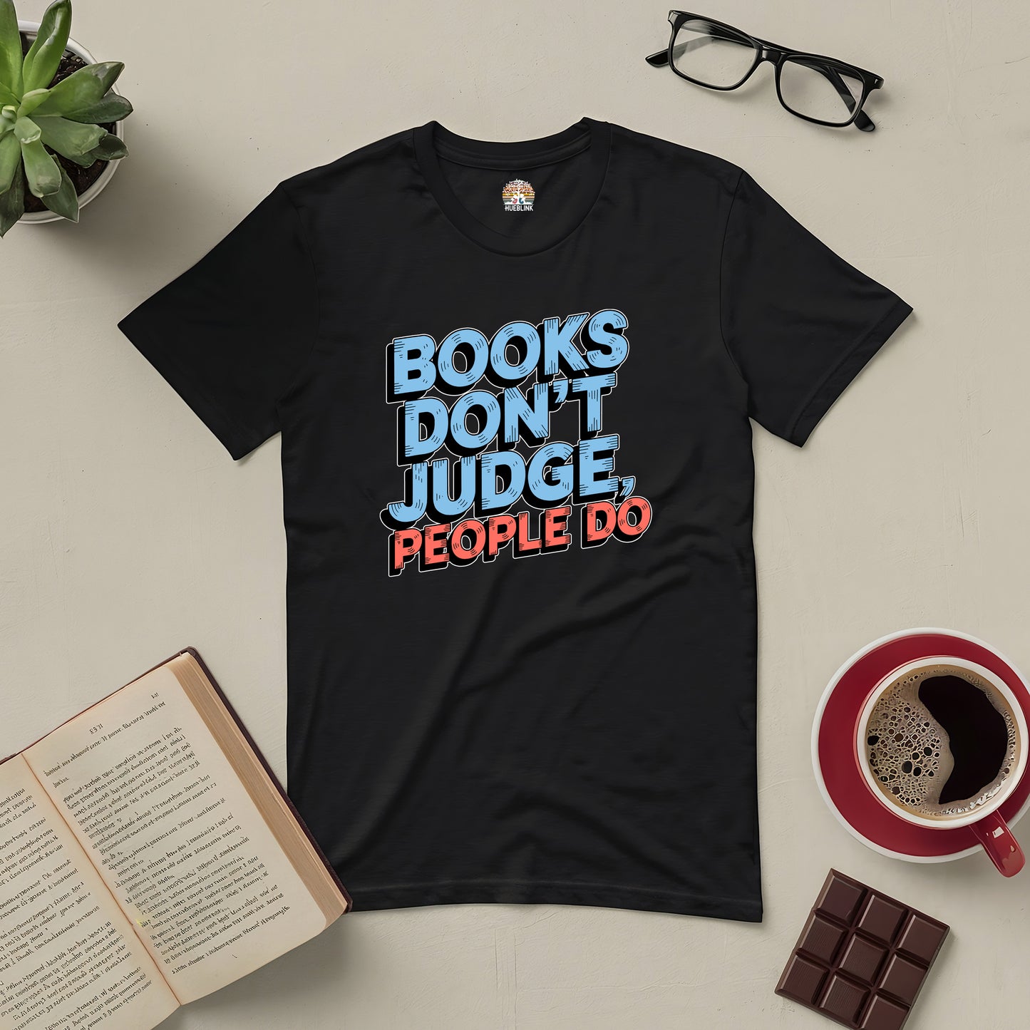 Black tee with "Books Don't Judge, People Do" slogan, surrounded by books, glasses, coffee, and chocolate on a beige surface.