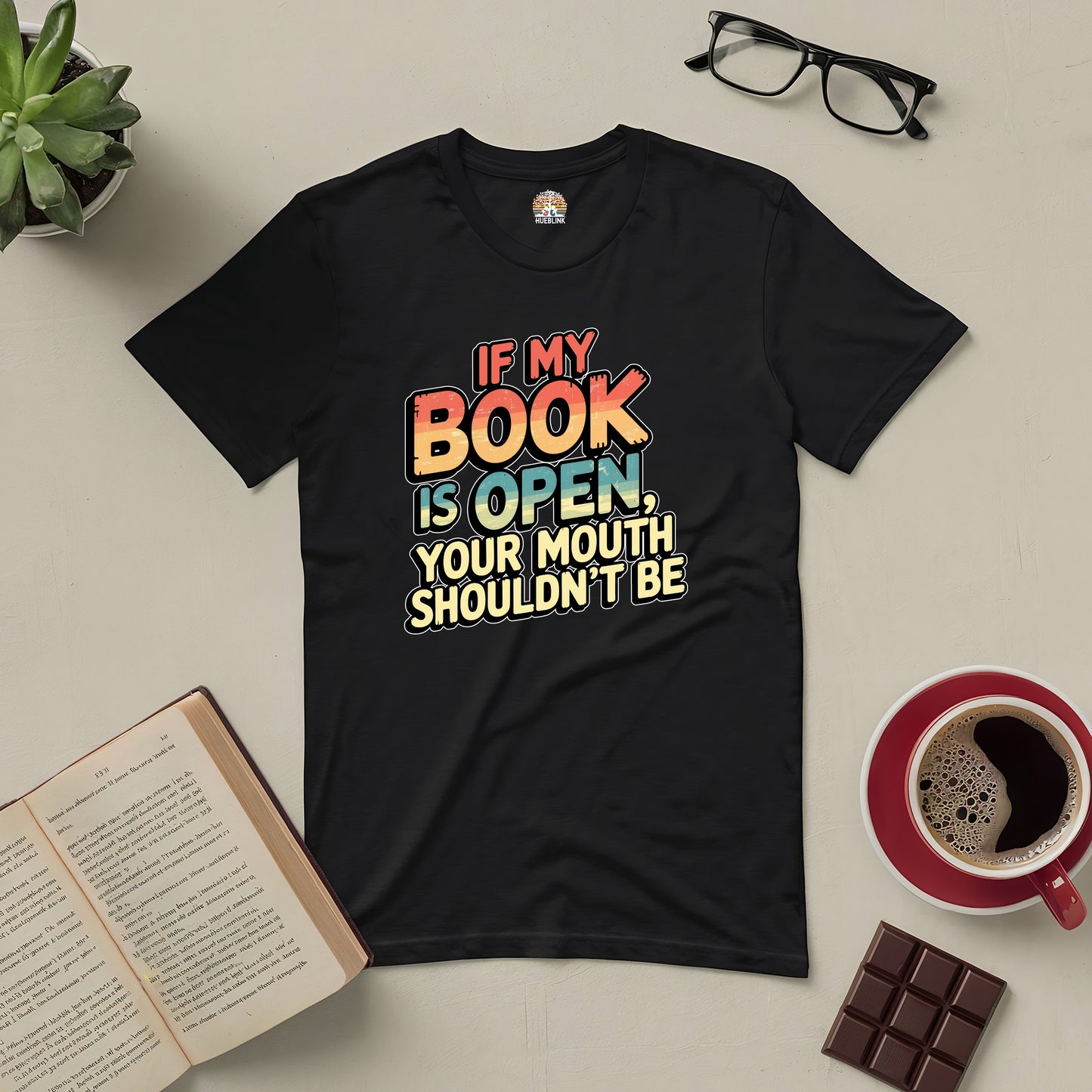 Black tee with "If My Book Is Open, Your Mouth Shouldn't Be" text, surrounded by open book, glasses, coffee, and chocolate.