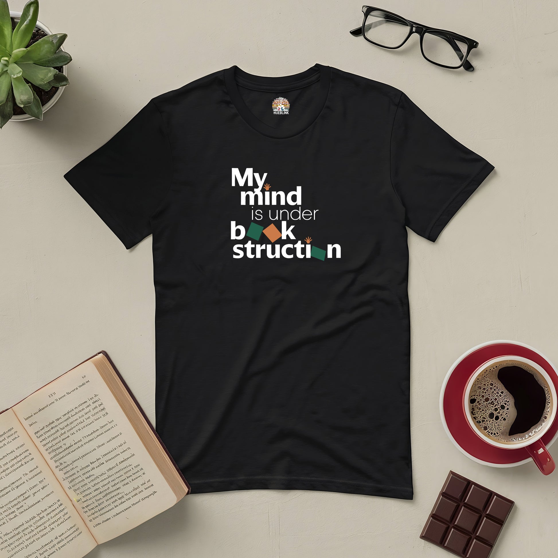 Black tee with "My Mind is Under Book-struction" text, surrounded by an open book, coffee cup, chocolate, and eyeglasses on a desk.