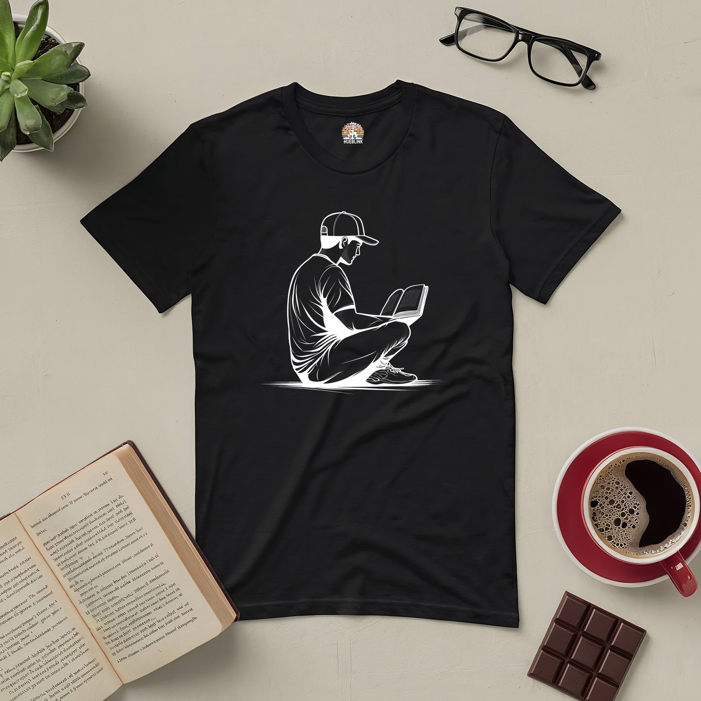 Black 'Reading in Serenity Tee' featuring a silhouette of a person reading, surrounded by books, coffee, and glasses on a table.