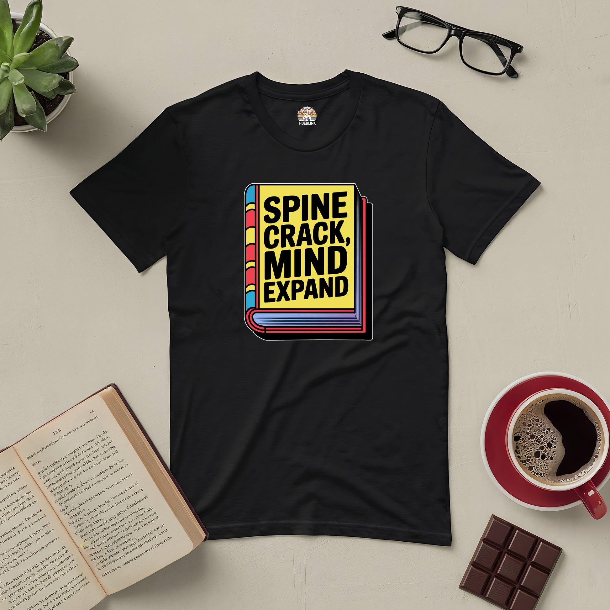 Black "Spine Crack, Mind Expand" tee laid flat, with open book, glasses, coffee, and chocolate beside it on a beige surface.