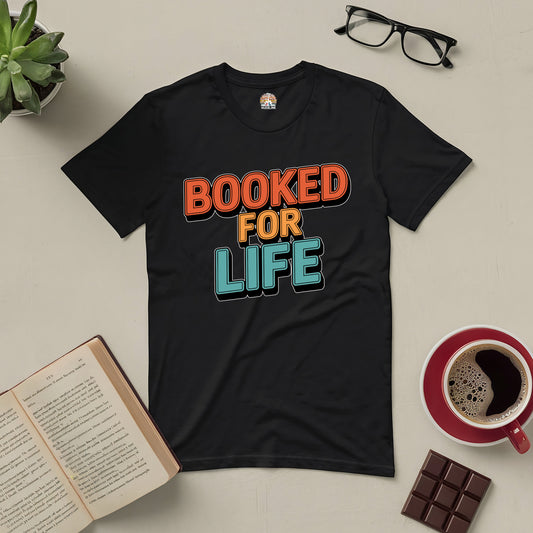 Black "Booked for Life" tee for book lovers, featuring bold colorful text on a flatlay with an open book, coffee, and chocolate squares.