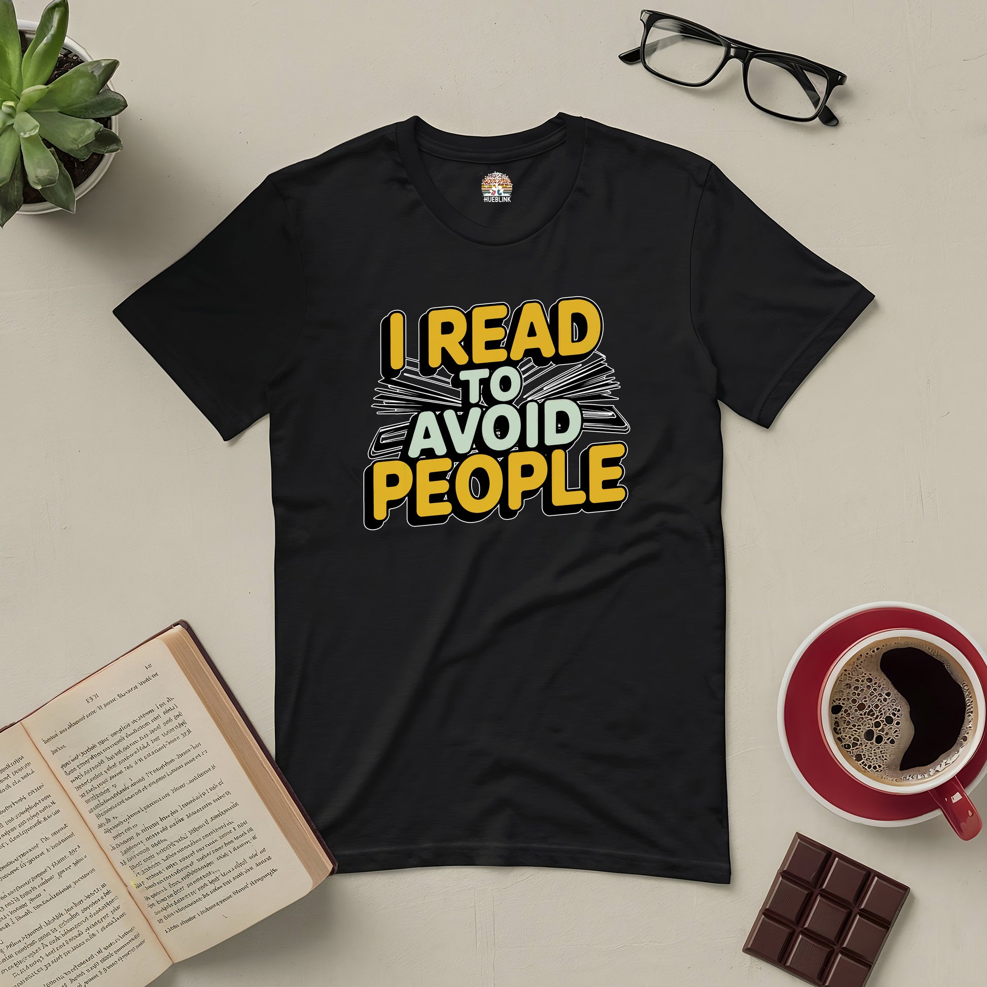 Black tee with "I Read To Avoid People" design, surrounded by books, glasses, coffee, and chocolate, ideal for book lovers.