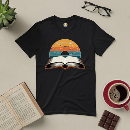 "Narratives in Color Tee featuring an open book under a vibrant sunset sky, symbolizing colorful reading journeys, surrounded by a cup of coffee and glasses."