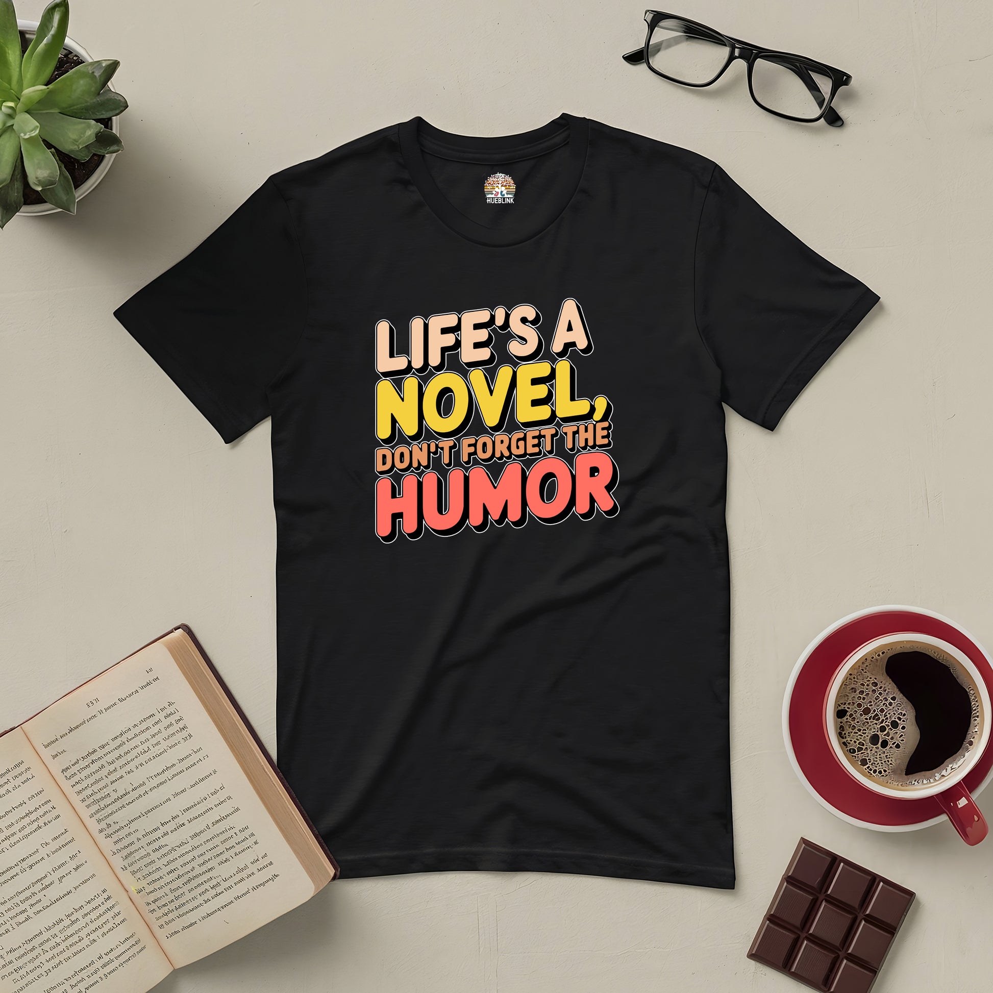 Black tee with "Life's a Novel, Don't Forget the Humor" text, surrounded by book, coffee, chocolate, and glasses on a table.