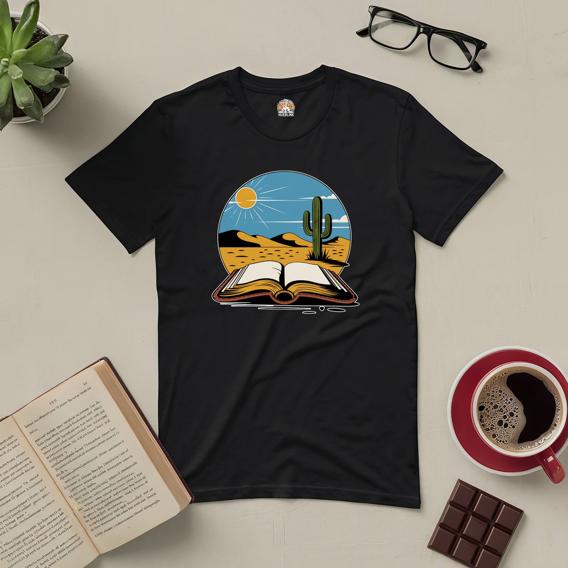 Black Silent Desert Tee with desert and book design, surrounded by an open book, glasses, plant, coffee cup, and chocolate.