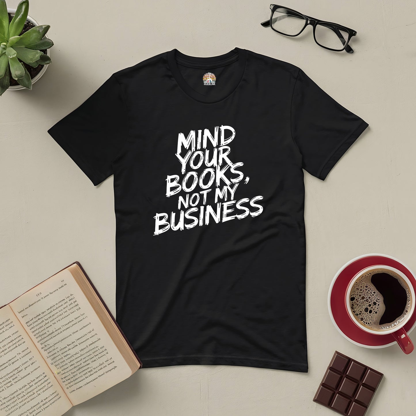 Black tee with "Mind Your Books, Not My Business" text, surrounded by an open book, coffee, glasses, and chocolate on a table.