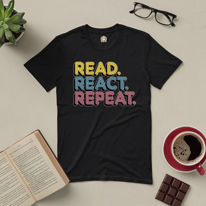 "Read React Repeat Tee with colorful text, surrounded by an open book, glasses, a red coffee cup, chocolate, and a small plant"