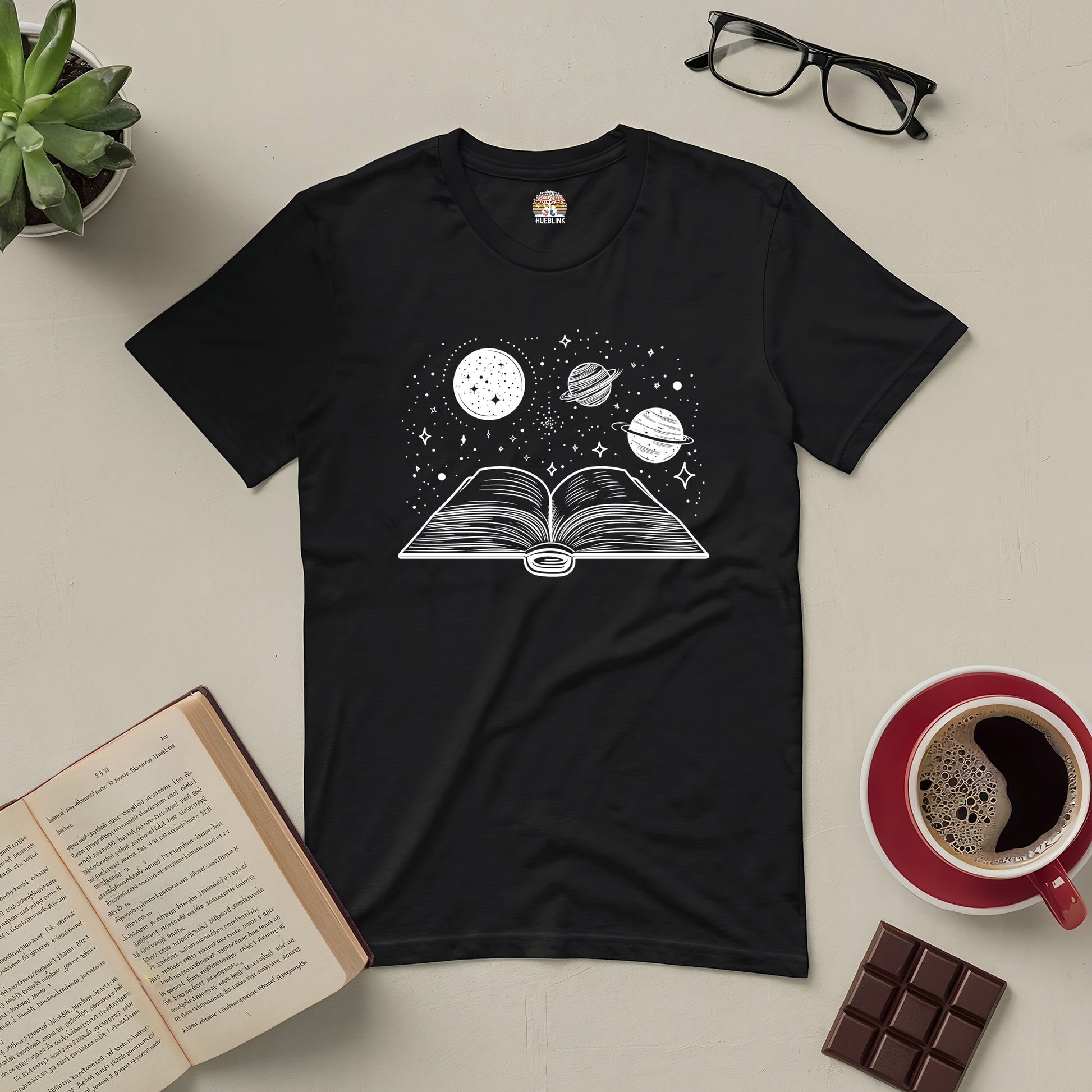 "Journey Through Space Tee with galaxy and book design, surrounded by open book, coffee, and chocolate on a table"