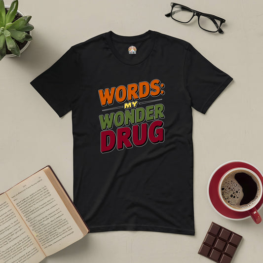 Black tee with "Words: My Wonder Drug" design, surrounded by book, coffee, glasses, plant, and chocolate on a light background.