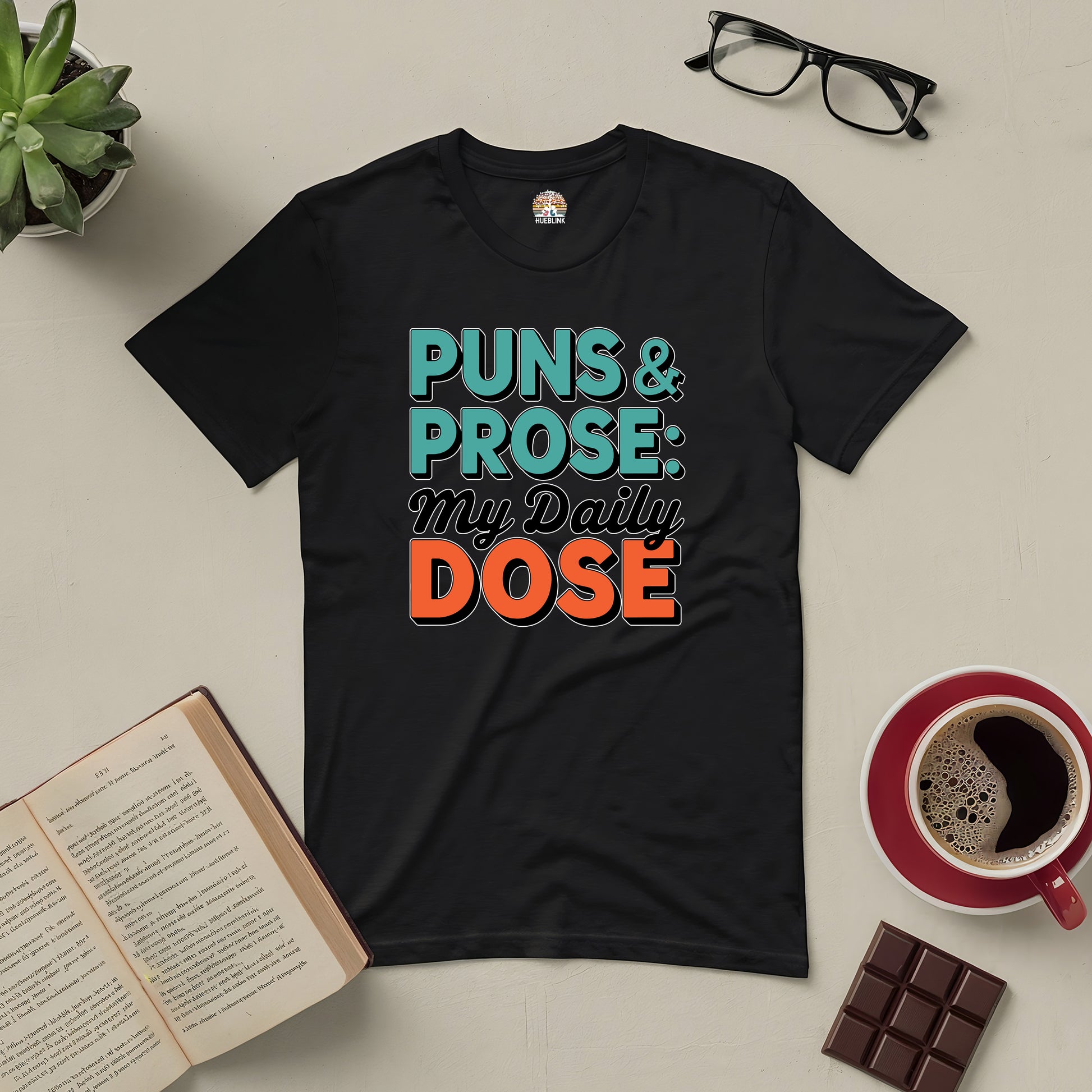 Black Puns & Prose Tee with witty text surrounded by book, coffee, glasses, plant, and chocolate bar.