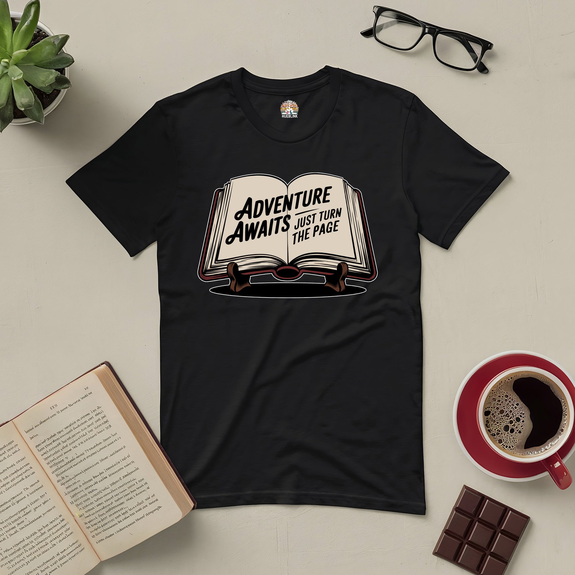 Black "Adventure Awaits: Just Turn the Page" tee on a table with books, glasses, coffee, chocolate, and a plant.