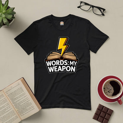 "Words: My Weapon Tee featuring bold design with lightning bolt and book is shown with coffee, glasses, chocolate, and an open book"