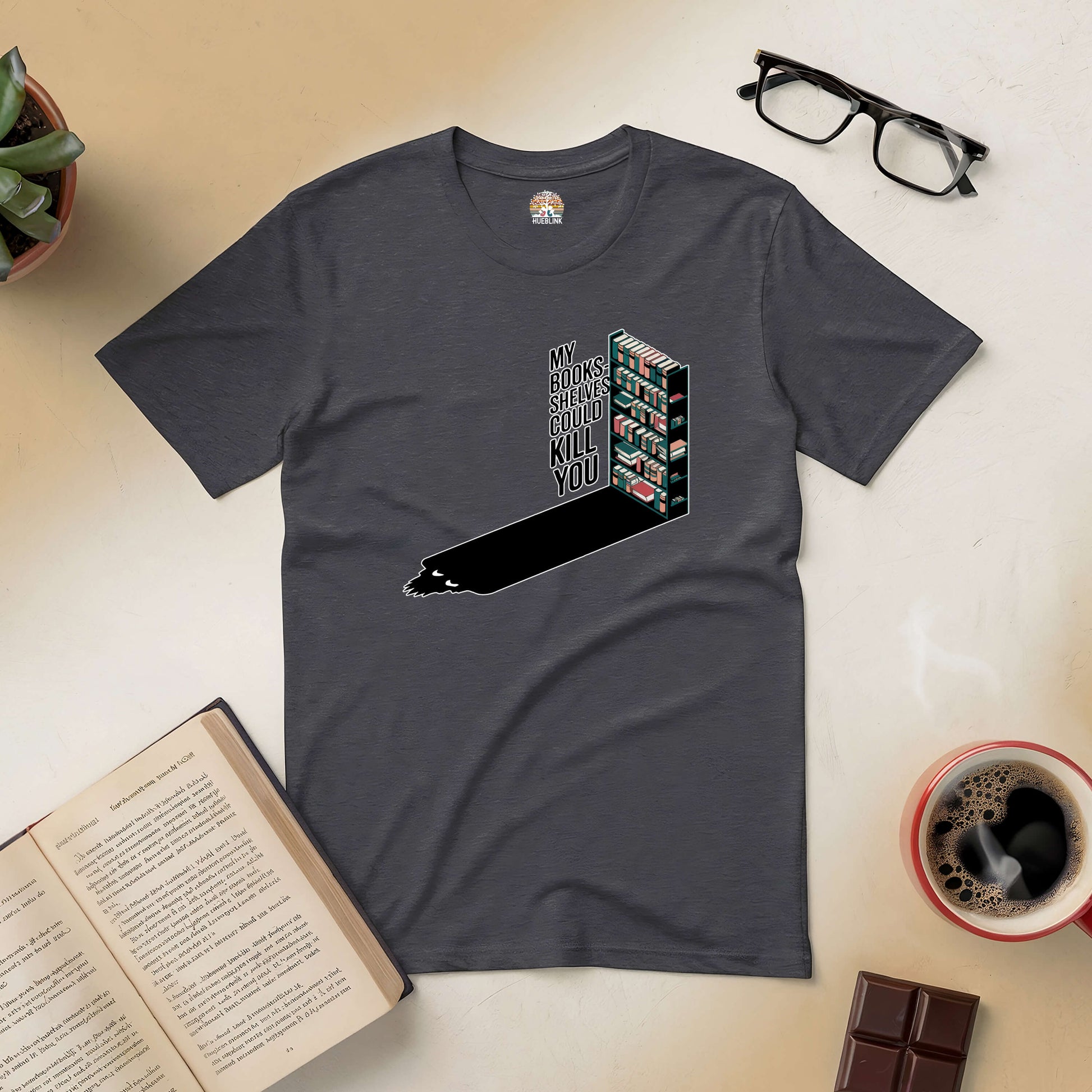 "Dark gray tee with 'My Bookshelves Could Kill You' graphic for book lovers with humor and edge, surrounded by coffee and open book."