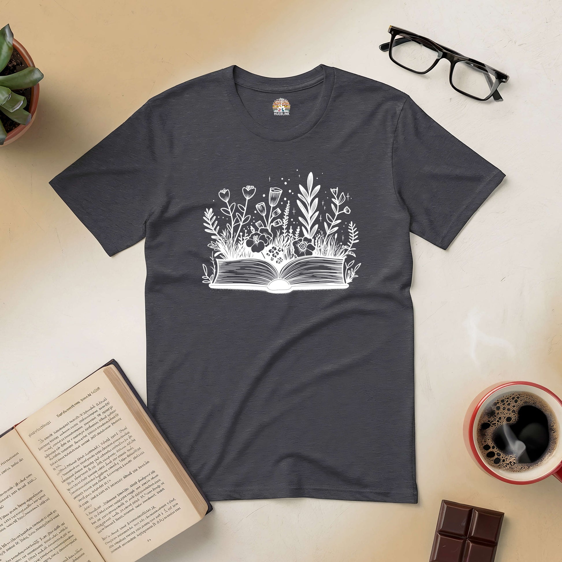 "Books and Blooms Tee with floral design, open book, surrounded by coffee, chocolate, glasses, and a plant, perfect for nature lovers"