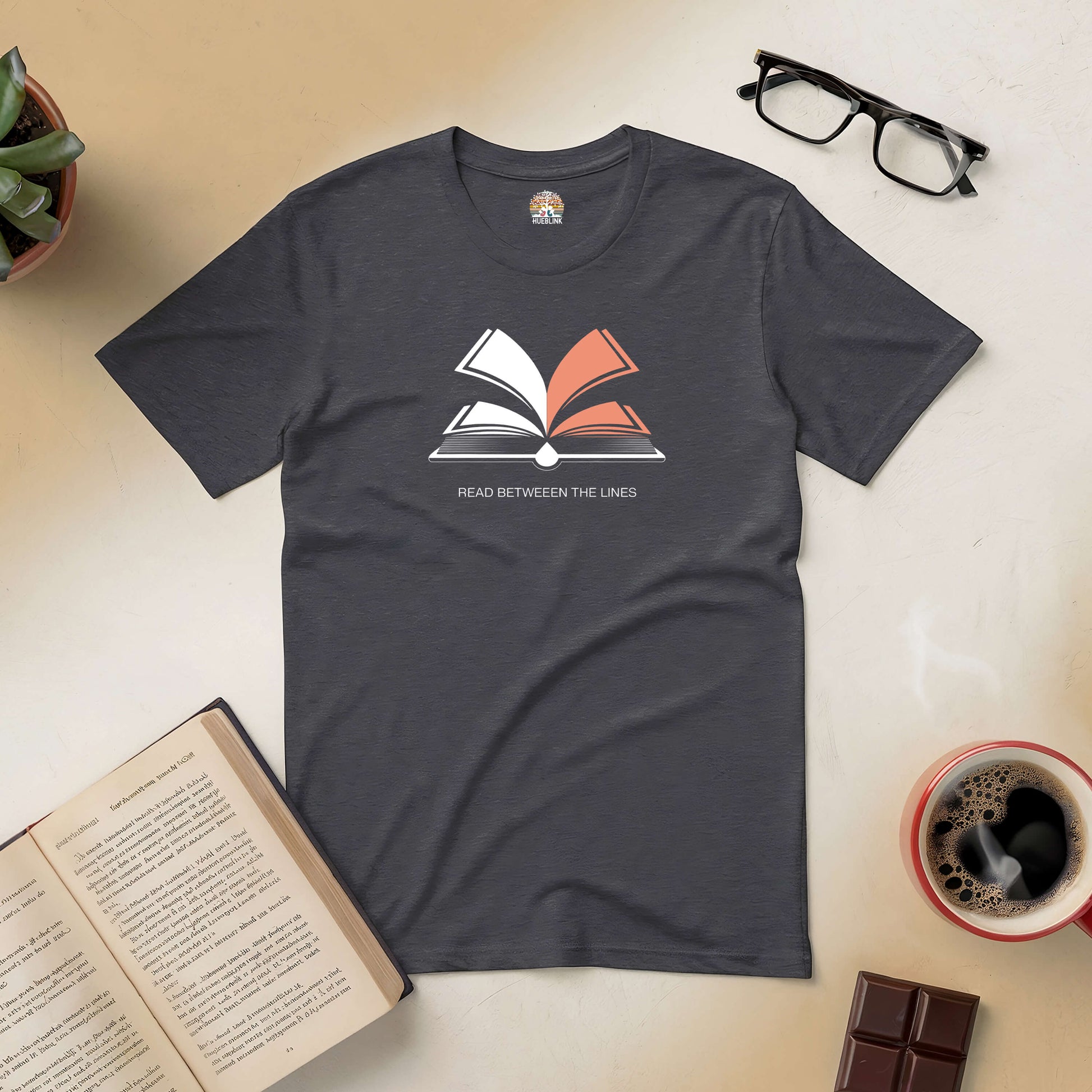 Dark grey "Read Between The Lines" tee with open book graphic, surrounded by glasses, coffee, chocolate, and a plant.