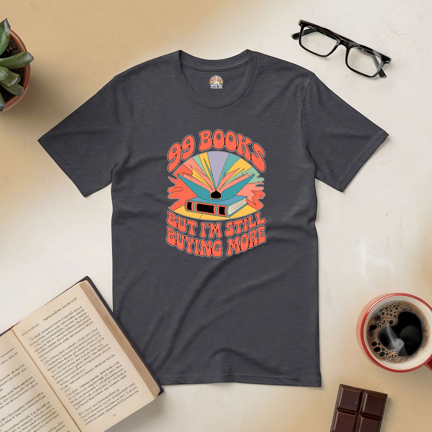 Charcoal tee with "99 Books But I'm Still Buying More" design, surrounded by a book, coffee, and glasses on a cozy reading setup.