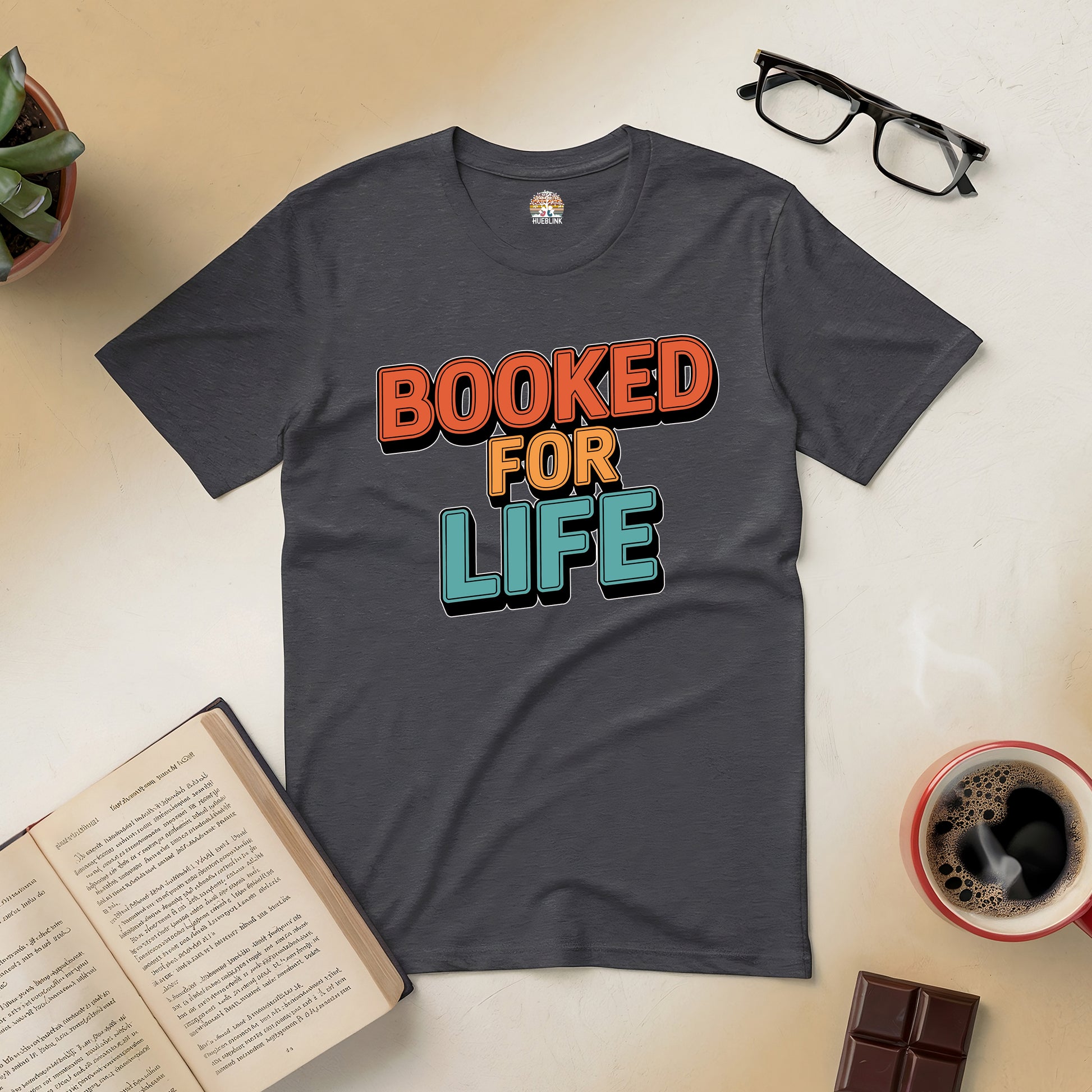 "Booked for Life Tee for devoted readers featuring bold text, surrounded by books, glasses, and coffee on a cozy table setting."