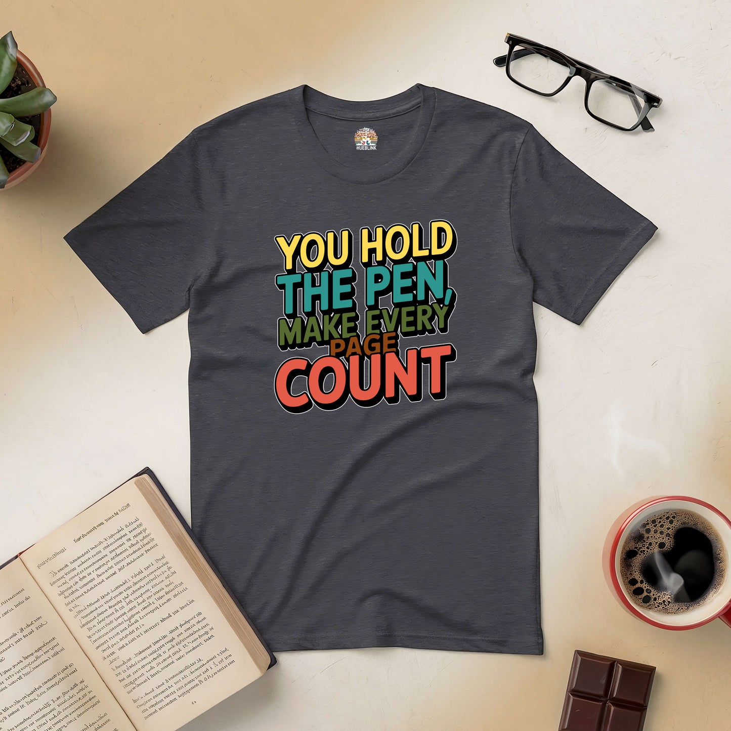 Inspirational "You Hold the Pen, Make Every Page Count" tee on table with book, coffee, glasses, plant, and chocolate.