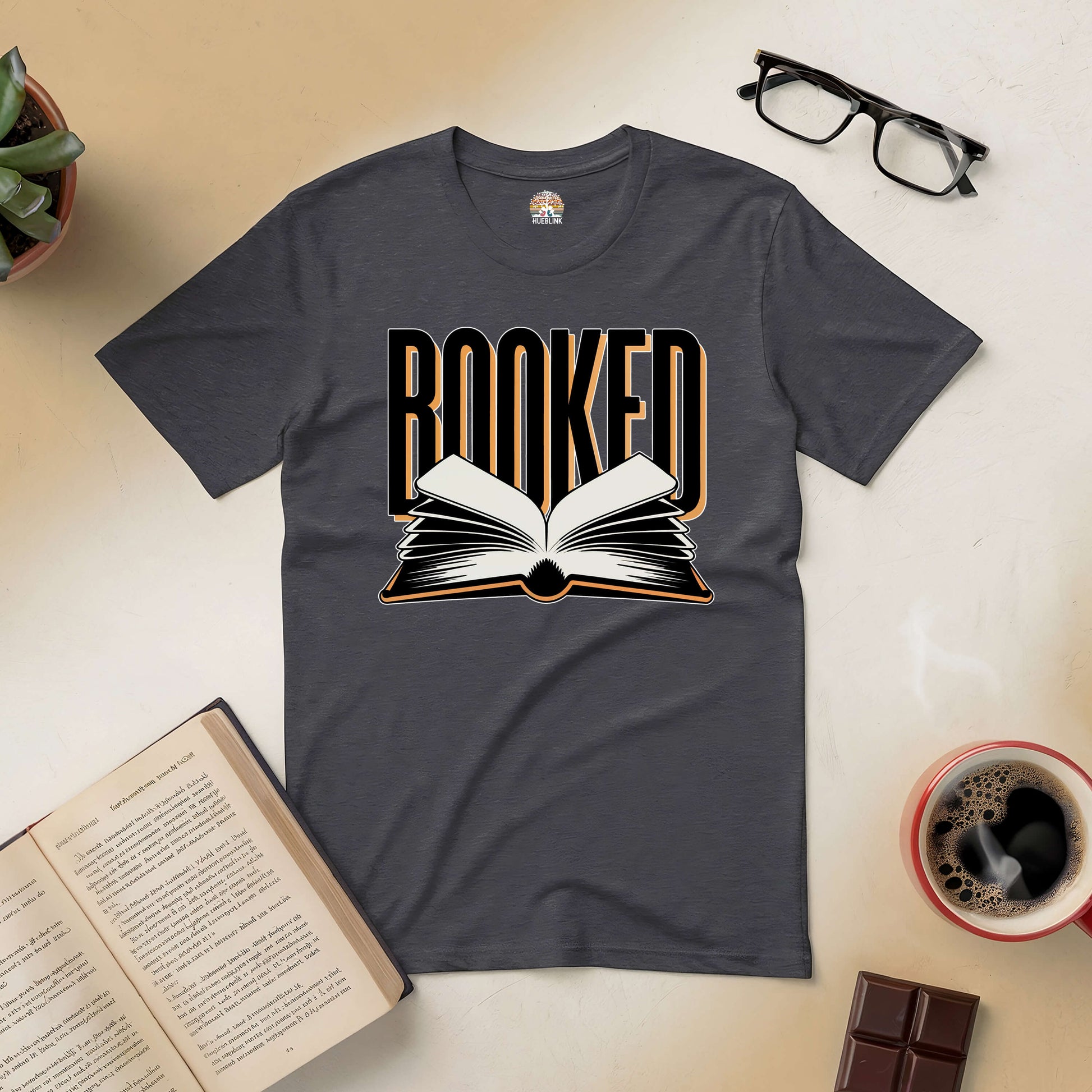 "Booked Tee for book lovers, featuring an open book graphic on a dark shirt, surrounded by coffee, chocolate, and open book."