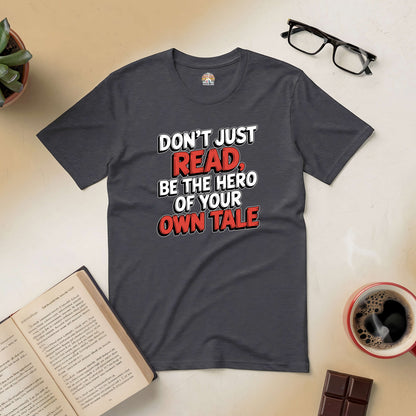 "Be the Hero of Your Own Tale Tee with motivational text on dark fabric, surrounded by book, coffee, and glasses."