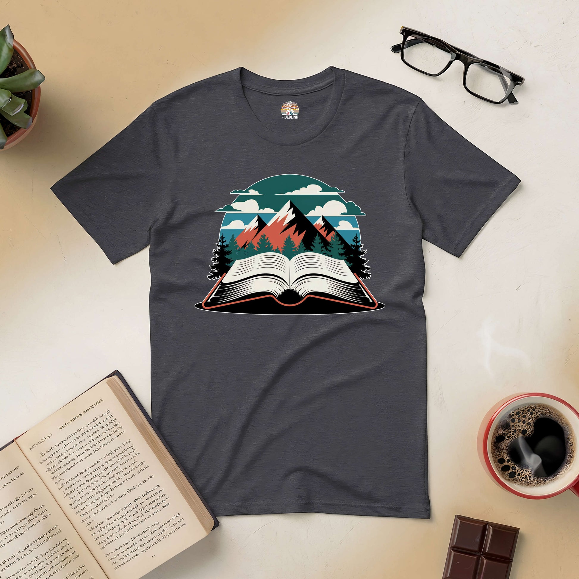 Graphic t-shirt with an open book and mountain design, surrounded by coffee, glasses, and an open book on a tabletop.