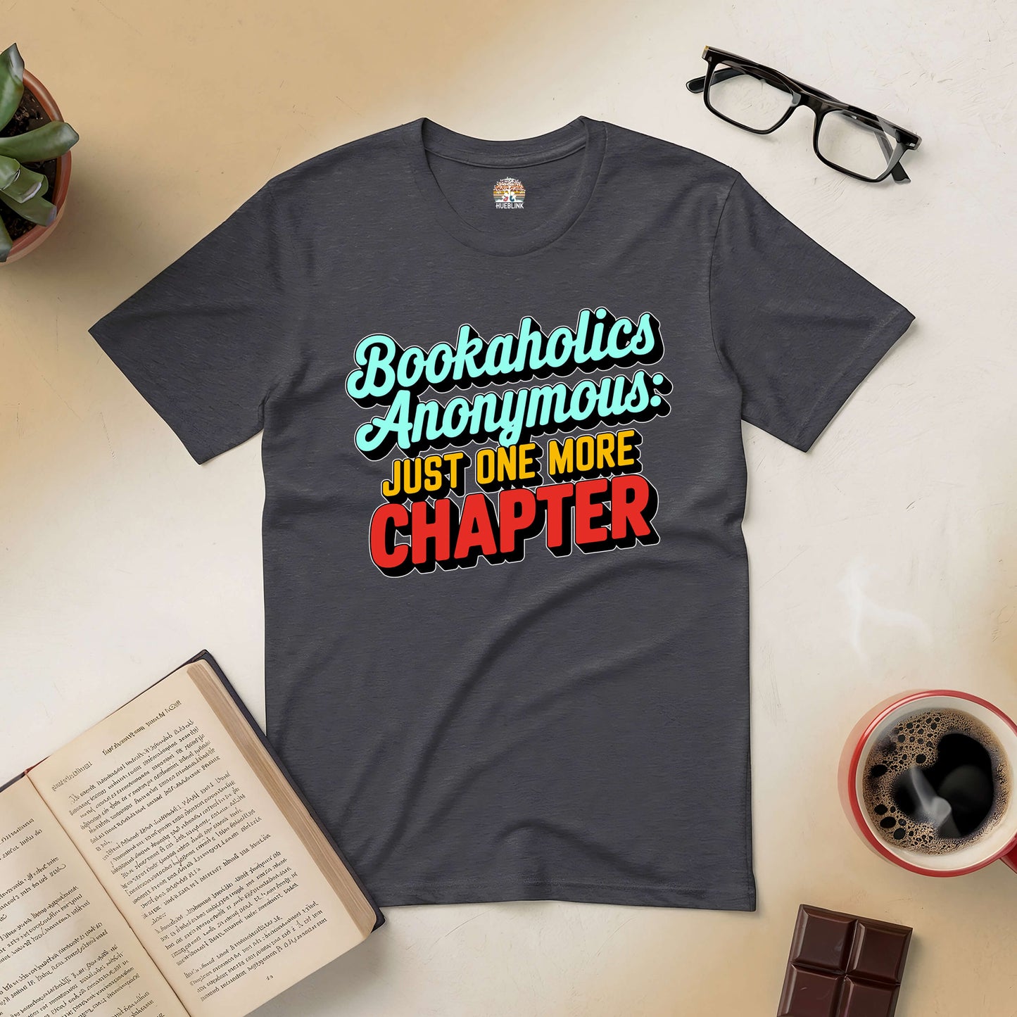 Bookaholics Anonymous Tee with "Just One More Chapter" text, surrounded by glasses, open book, and coffee cup. Perfect for avid readers.