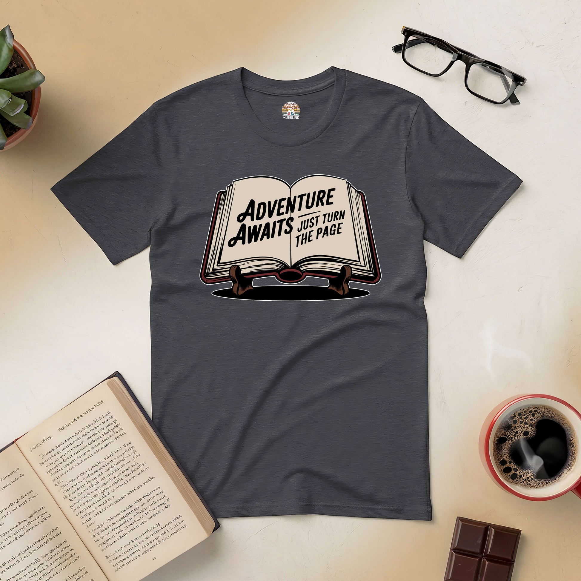 Adventure Awaits tee with open book graphic, coffee, glasses, and chocolate on a table.