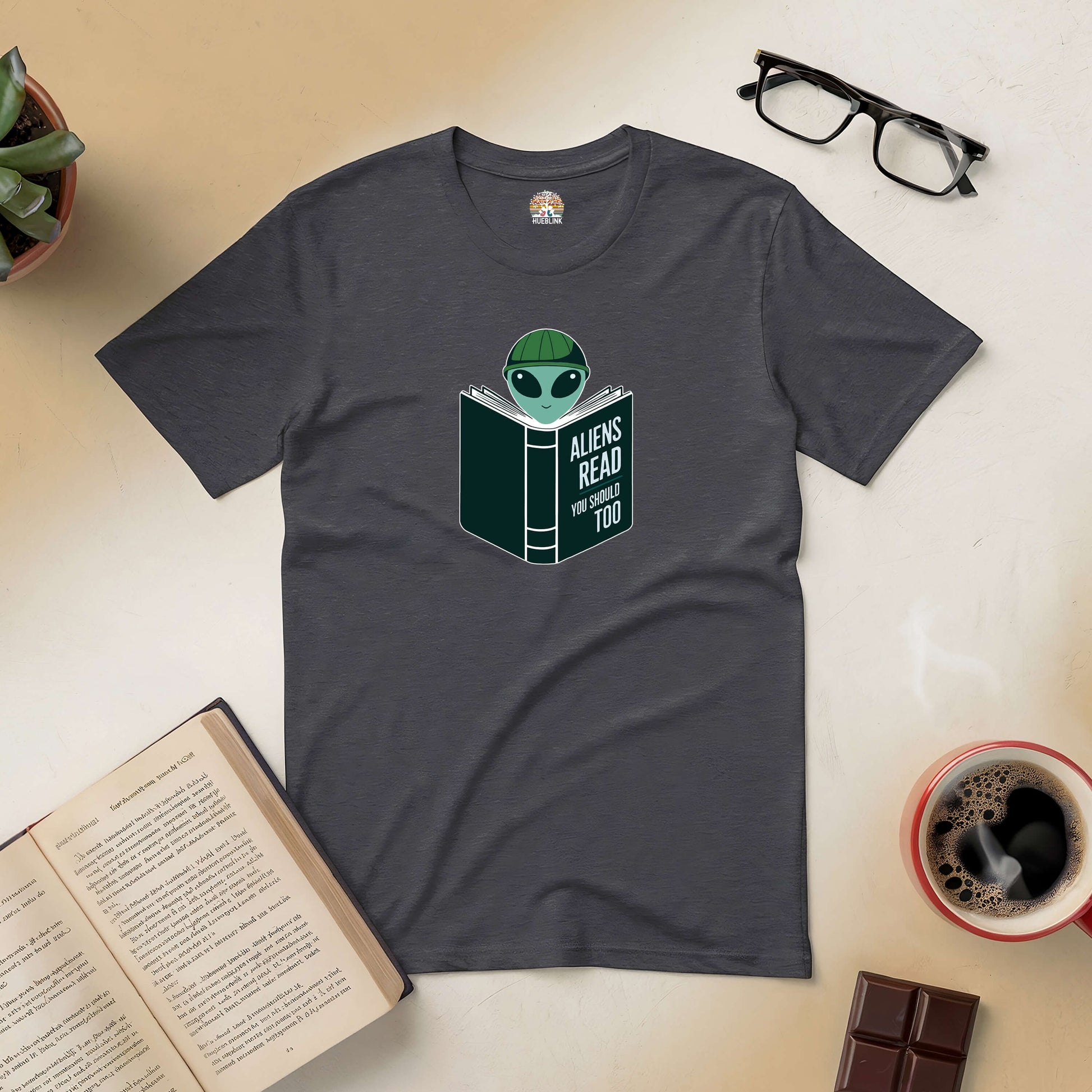 Grey "Intergalactic Habit" tee featuring alien reading a book, surrounded by coffee, chocolate, and open book on a table.