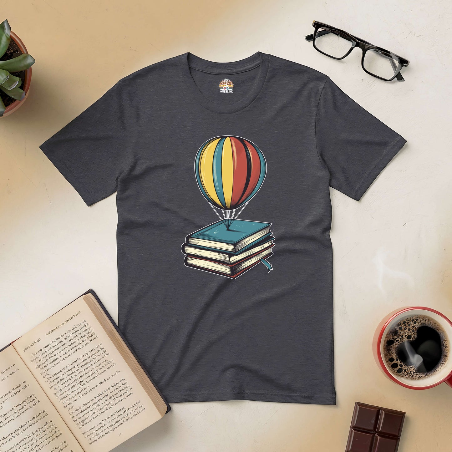 Passport to Dreamland tee with hot air balloon and books, surrounded by open book, coffee, and glasses on a light background.