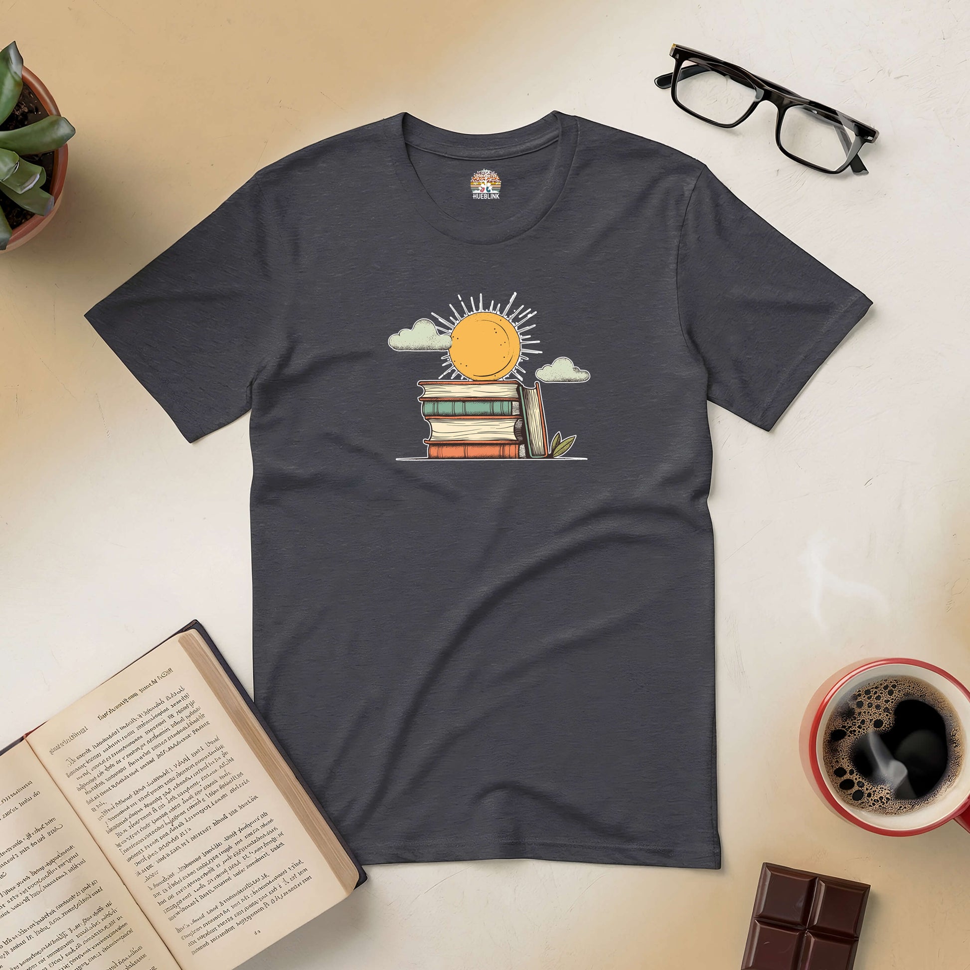 "Literary Sunshine Tee with sun and books design for book lovers, surrounded by open book, glasses, plant, coffee, and chocolate"