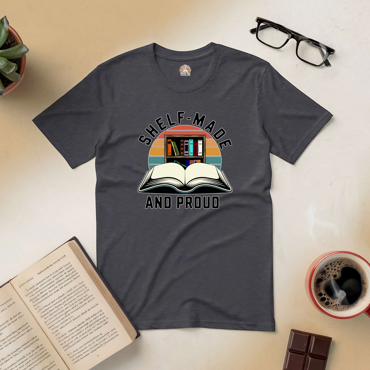 "Shelf-Made And Proud Tee with book graphic, surrounded by open book, glasses, coffee, and chocolate on a table"