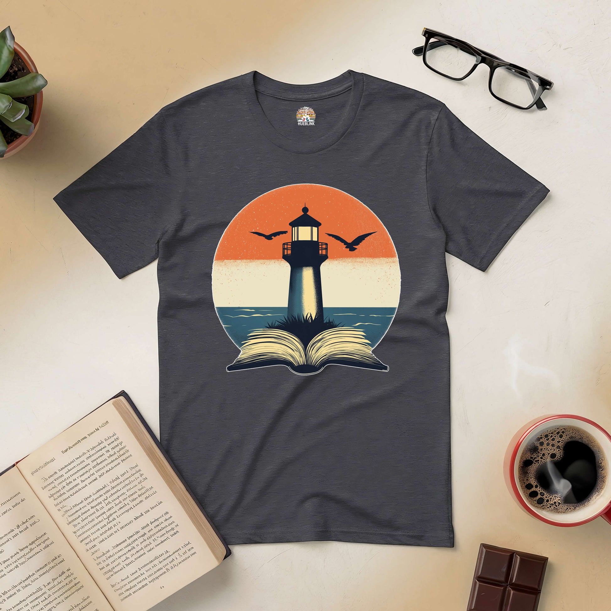 Charcoal t-shirt with a lighthouse and open book design, symbolizing exploration and wisdom, surrounded by books and coffee.